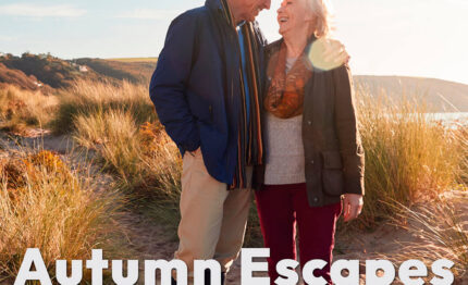 Elderly couple enjoying a coastal walk among sandy dunes during autumn, with sunlit grassy hills in the background.