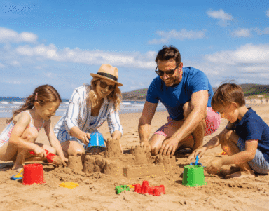Family building sandcastles on sunny beach by seaside hotel with golden sands, clear skies, and ocean views.