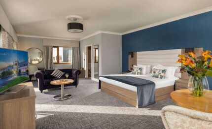 Spacious hotel suite with cosy king-size bed, elegant sofa, flat-screen TV, and vibrant flowers, offering a relaxing stay.