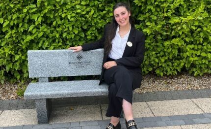 Smiling woman in professional attire on a bench, lush green garden backdrop, inviting hotel ambience.