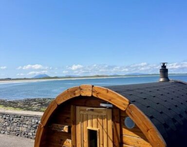 Cosy wooden sauna overlooking serene sea and coastline under clear blue sky at scenic hotel location.