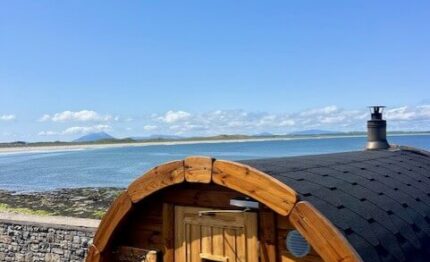 Cosy wooden sauna overlooking serene sea and coastline under clear blue sky at scenic hotel location.