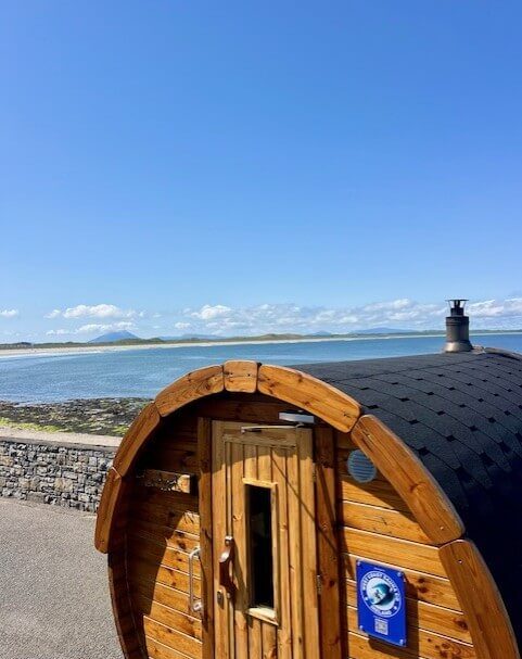 Cosy wooden sauna overlooking serene sea and coastline under clear blue sky at scenic hotel location.